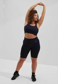 Mørkeblå ribbestrikket sportsbh og matchende høyt midjebykeshorts, begge laget av glatt, elastisk stoff, vist på en modell i en strekkeposisjon.