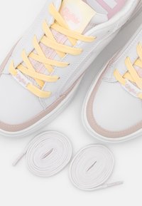 Baskets blanches avec des lacets jaunes, des accents roses et une texture en cuir lisse. Comprend deux lacets blancs de rechange.