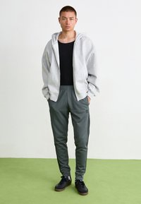 Pelēks zip-up džemperis virs melnas bezmuguras blūzes, apvienots ar zaļām sporta biksēm ar melnām svītrām un logo, kā arī melniem sporta apaviem ar gumijas zoli.