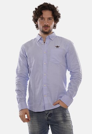 Camisa de botón de color azul claro en tejido de algodón con mangas largas, que presenta un bolsillo en el pecho y un logotipo bordado encima del bolsillo.