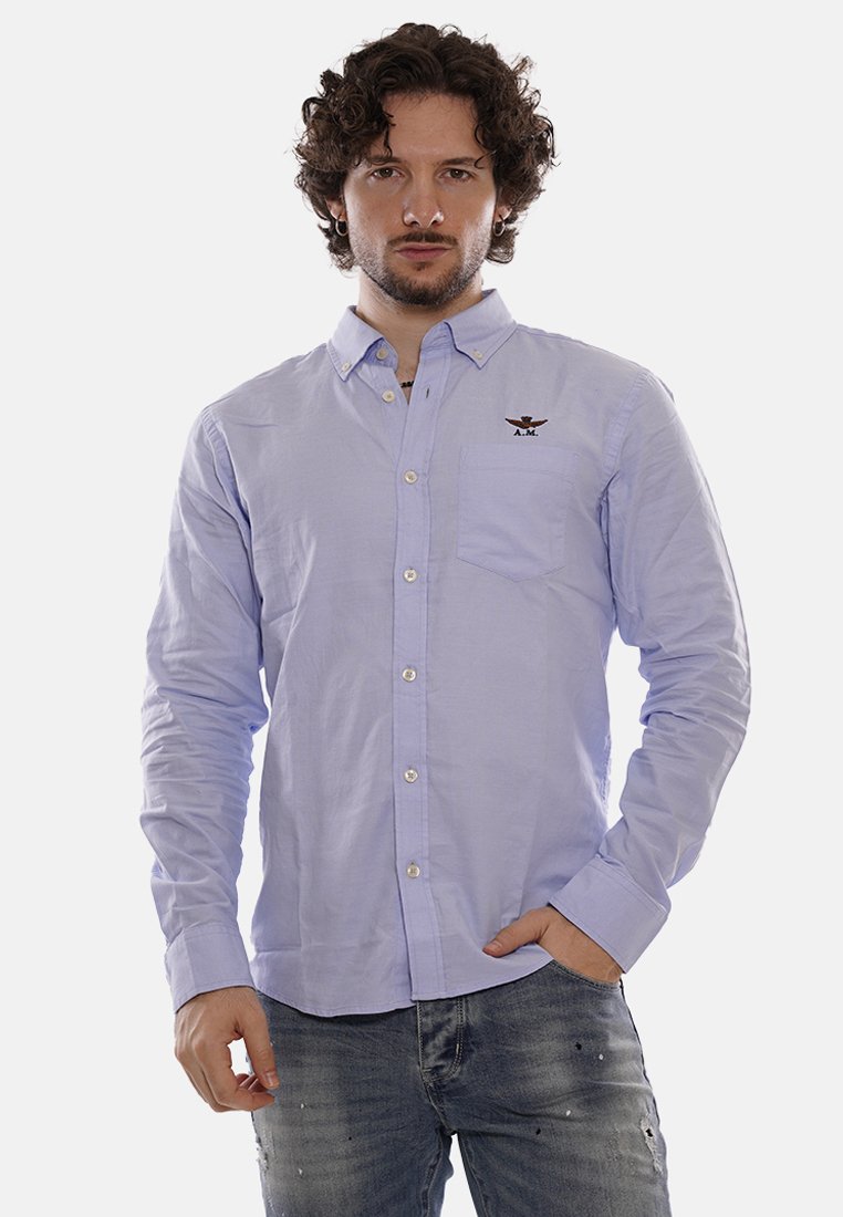 Camisa de botón de color azul claro en tejido de algodón con mangas largas, que presenta un bolsillo en el pecho y un logotipo bordado encima del bolsillo.