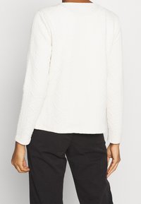 Weißer strukturierter Pullover mit Zopfmuster, langen Ärmeln und geradem Saum, kombiniert mit schwarzen Hosen.