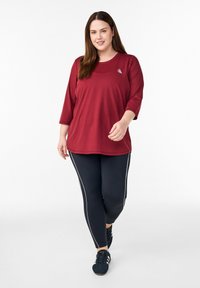 Rotes sportliches Oberteil mit Dreiviertelärmeln, kombiniert mit schwarzen Leggings mit weißen Seitenstreifen und schwarzem Schuhwerk. Glatte Stoffstruktur.