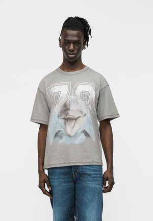 Young man with braided hair wears gray t-shirt featuring large 78 and graphic of a smiling dolphin, paired with blue jeans, standing against white background.