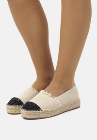 Espadrillas in tela beige con bordi sfilacciati e punta nera glitter, dotate di suola in juta intrecciata. Design casual e leggero.