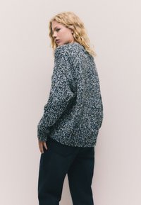 Jersey de punto en tonos negro, blanco y azul. Textura gruesa, ajuste holgado, escote redondo y puños acanalados, combinado con pantalones oscuros.