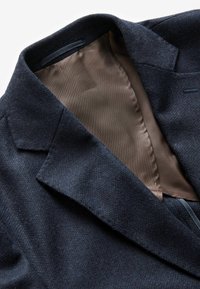 Blazer en laine marine avec un extérieur texturé, un intérieur en satin marron, des revers à cran et un détail de poche poitrine unique.
