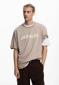 Beige oversized T-shirt med "DESIGUAL" printet i hvid, korte ærmer, lagd over en hvid skjorte. Brune bukser med sidelommer.