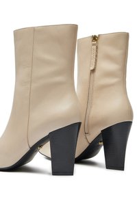 Kremas odiniai kulkšnies ilgio batai su lygiu paviršiumi, kontrastingais juodais blokiniu kulnais ir aukso spalvos užtrauktuko detalė gale.