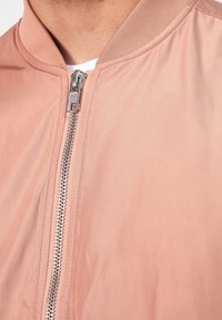 Jasnoróżowa kurtka bomberka z gładkiego materiału, z prążkowanym kołnierzem i srebrnym zamkiem błyskawicznym z przodu. Prosty, minimalistyczny design.