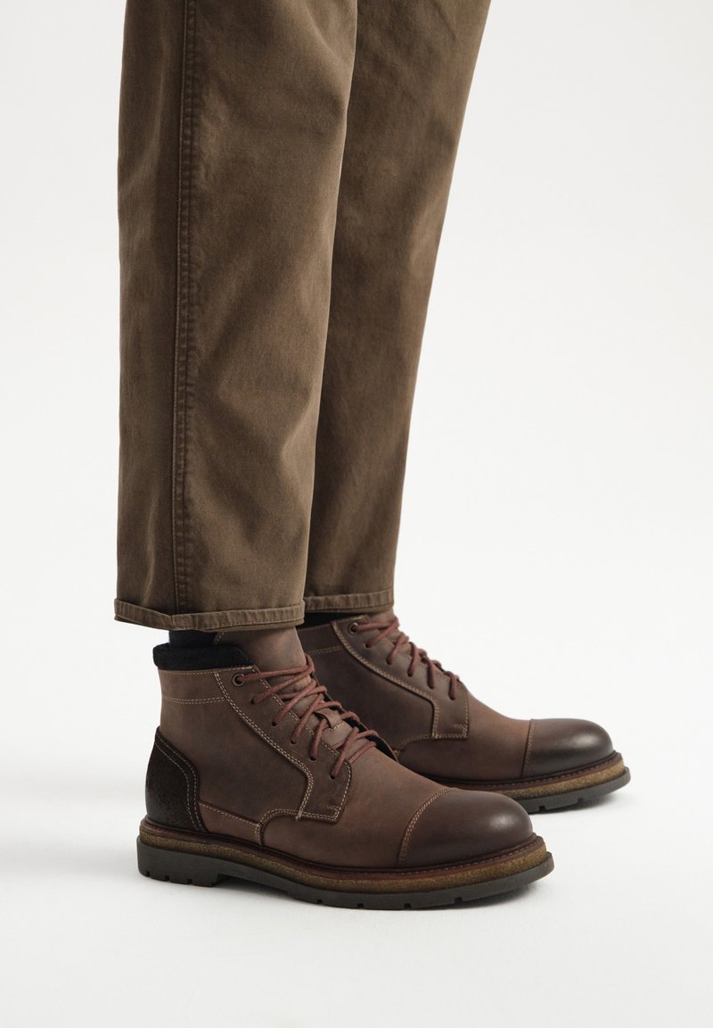 Brązowe skórzane botki za kostkę z okrągłym noskiem, kontrastującą ciemną częścią kostki i ciemną gumową podeszwą. Noszone z podwiniętymi brązowymi spodniami.