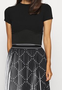 Czarny prążkowany top z okrągłym dekoltem, zestawiony z plisowaną czarną spódnicą z powtarzającym się białym wzorem logo „GUESS”.