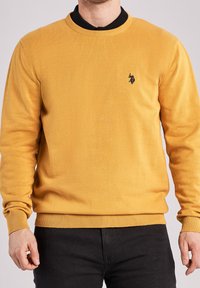 Senapsgul stickad tröja med rund halsringning och ribbade ärmslut och nederkant. Har en liten svart logotyp på bröstet. Enkel design.