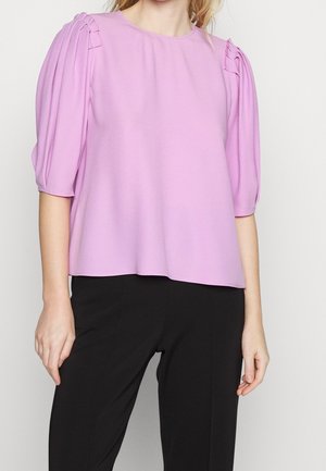 Kvinde iført en lilla bluse med korte ærmer og plisserede skulderdetaljer sammen med sorte bukser, stående foran en ensfarvet baggrund.