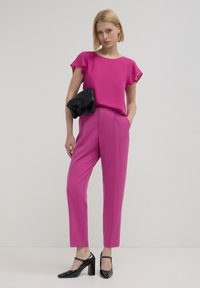 Blusa fucsia a maniche corte abbinata a pantaloni fucsia sartoriali. La modella tiene una borsa a mano nera con texture. Scarpe nere con tacco completano il look.