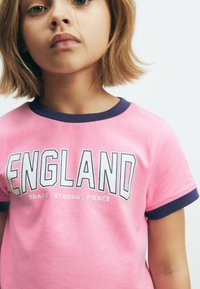 Vaaleanpunainen puuvillapaita, jossa on pyöreä tummansinisä kaulus. Paidassa lukee isoilla valkoisilla kirjaimilla "ENGLAND" ja pienemmällä tekstillä "BRAVE. STRONG. FIERCE."