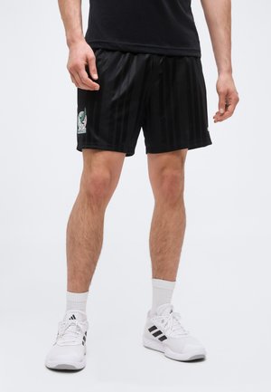 Mann trägt schwarze Sportshorts mit Mexiko-Logo, weiße Sportsocken und weiße Adidas-Sneakers, steht vor einem einfarbigen Hintergrund.