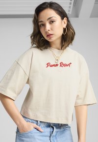 Beige bomulds crop top med korte ærmer, der har røde broderede "Puma Resort"-tekster og et lille Puma-logo, parret med lyseblå jeans.