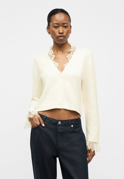 Γυναίκα που φοράει ένα κρεμ ζακέτα με κουμπιά και δαντέλα στο γιακά και τα μανίκια, σε συνδυασμό με σκουρόχρωμα ψηλόμεσα τζιν.