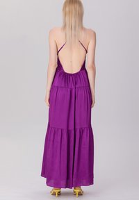 Robe maxi violette avec un dos décolleté, présentant un design à volants et des fines bretelles. Tissu doux et forme fluide. Portée avec des talons jaunes.
