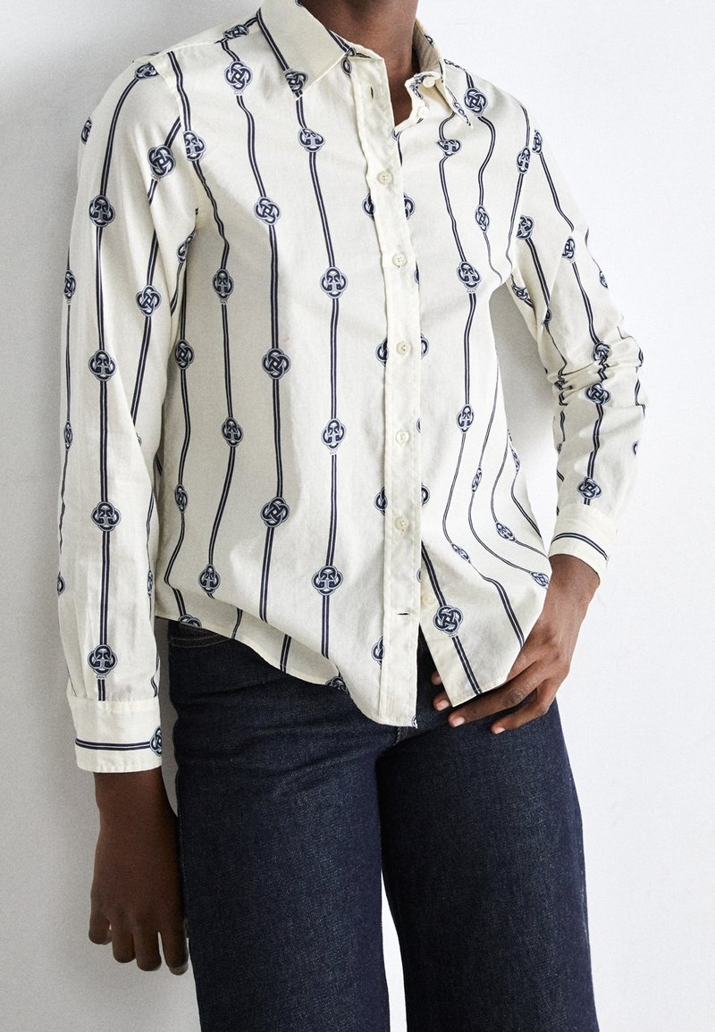 Person, die ein weißes langärmliges Hemd mit schwarzen geometrischen vertikalen Streifen und dunkelblauen Jeans trägt, vor einem schlichten Hintergrund.