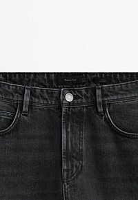 Jean noir slim avec fermeture à boutons, poches devant, passants pour ceinture et une étiquette de marque visible à l'intérieur de la ceinture.