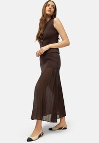 Abito marrone senza maniche con parte superiore aderente e gonna plissettata, con dettagli in bottone dorato. Abbinato a scarpe slingback beige e nere.