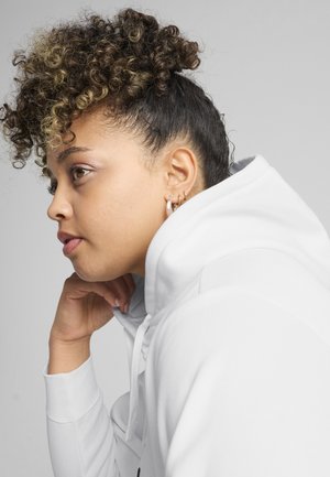 Jong persoon met krullend haar in een witte hoodie, met de kin op de hand terwijl die vooruit kijkt tegen een effen grijze achtergrond.