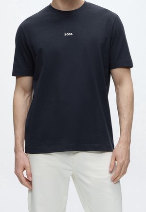 Homme portant un T-shirt noir uni à manches courtes avec le texte "BOSS" sur la poitrine, associé à un pantalon clair.