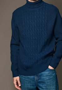 Homme portant un pull à col roulé en tricot côtelé bleu marine et un jean bleu, une main dans la poche, devant un fond marron uni.