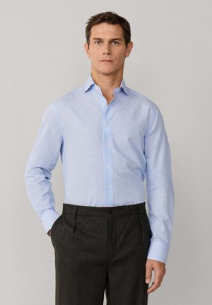 Homme portant une chemise bleu clair à boutons, rentrée dans un pantalon gris foncé, debout avec une main dans la poche devant un fond uni.