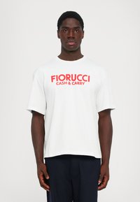 Jauns vīrietis, tērpies baltā T-kreklā ar sarkano tekstu "FIORUCCI CASH & CARRY", stāv pretī vienkāršam gaišam fona.