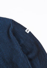 Artimu planu matoma juoda ir mėlyna juostuota megztinė medžiaga su balta stačiakampio formos etikete, siuvama ant krašto.