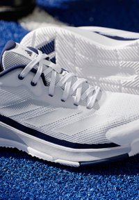 Bílé sportovní boty s texturovanou síťovinou, námořnickými akcenty a tkani�čkami, s odolnou podešví s gripovým vzorem. Umístěno na modré umělé trávě.