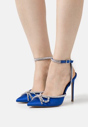 Des chaussures à talons hauts en satin bleu avec des orteils pointus, ornées d'un nœud embellie de cristaux et d'une bride de cheville, présentant une finition métallique texturée.