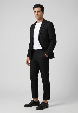 Man staat, gekleed in een zwart geruit pak, wit t-shirt en zwarte nette schoenen tegen een effen grijze achtergrond.