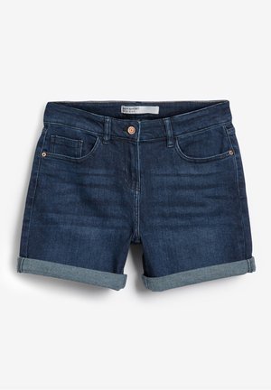 Jeune femme portant un crop top blanc à pois, un cardigan vert menthe et un short en jean foncé retroussé, debout sur un chemin en bois à la plage.