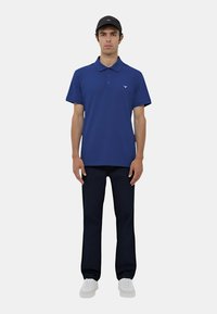 Polo shirt blu navy con colletto, maniche corte e logo sul petto; abbinato a pantaloni in denim scuri e sneakers bianche.