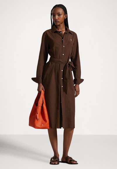 Woman wearing a brown buttoned midi dress with a waist tie, holding an orange leather handbag, and wearing brown sandals, standing on a plain background.