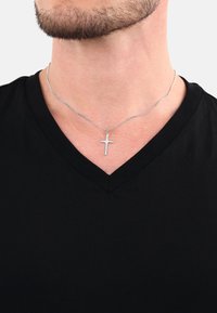 Silbernes Kreuzhalsband an einer dünnen Kette, getragen über einem schwarzen V-Ausschnitt-T-Shirt, vor einem neutralen Hintergrund.