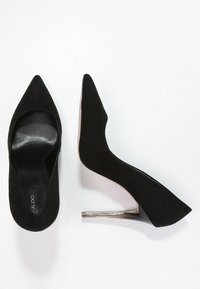 Svarta mockaskor med hög klack och spetsiga tår, där den ena visar innersulan och den andra har en metallisk stilettklack.