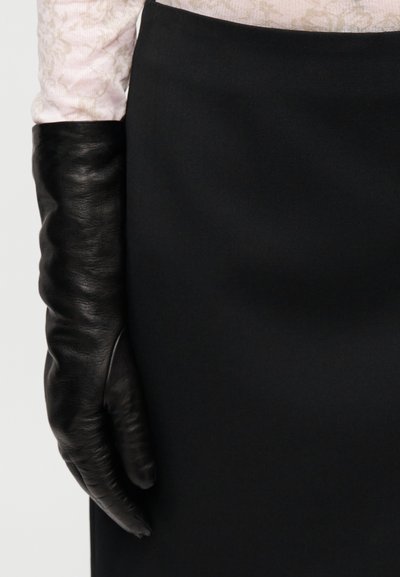 Gant en cuir noir avec une finition texturée, présentant une coupe ajustée. Porté sur une jupe noire unie, partiellement caché par un chemisier à motif.