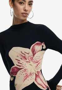 Ribba, pitkähihaine mekko laivastonsinisellä taustalla, jossa on suuri vaaleanpunainen ja kerma-valkoinen liljakuvio yhdellä puolella. Mekossa on istuva siluetti ja korkea kaulus.