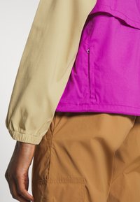 Personne portant un haut beige à manches longues, une veste violette avec une poche latérale et un pantalon marron avec une poche sur le côté.