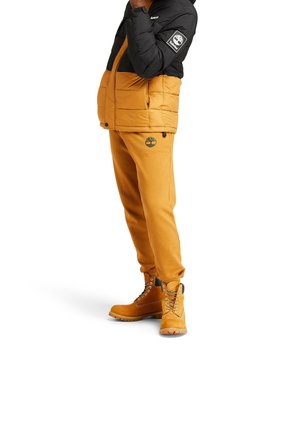 Żółta kurtka puchowa z czarnym górnym fragmentem, czarne logo na rękawie; pasujące żółte spodnie dresowe z logo na udzie; beżowe buty z sznurowadłami.