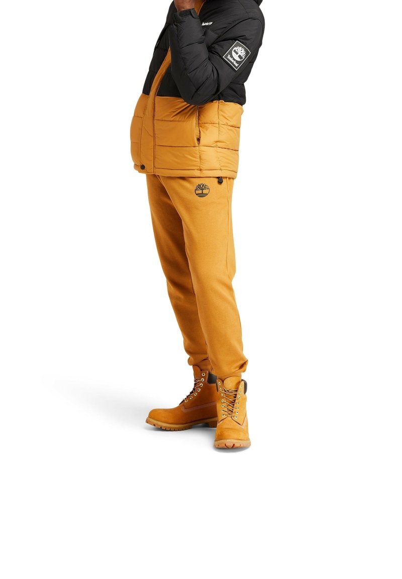 Żółta kurtka puchowa z czarnym górnym fragmentem, czarne logo na rękawie; pasujące żółte spodnie dresowe z logo na udzie; beżowe buty z sznurowadłami.