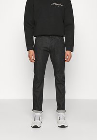 Person som bär svart långärmad tröja, mörka uppvikta jeans och vita sneakers, stående mot en enfärgad vit bakgrund.