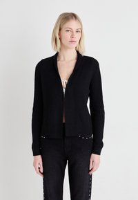 Cardigan negru cu nervuri, cu guler tip șal, mâneci lungi și design deschis în față, purtat peste un top deschis la culoare și asortat cu blugi închiși.