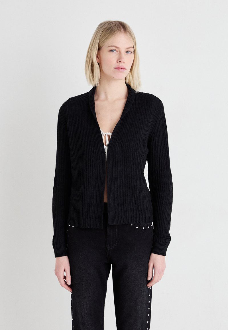 Cardigan negru cu nervuri, cu guler tip șal, mâneci lungi și design deschis în față, purtat peste un top deschis la culoare și asortat cu blugi închiși.