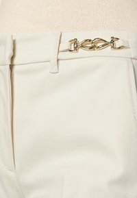 Pantalon blanc en tissu lisse et texturé. Présente un accent de chaîne dorée au niveau de la ceinture, avec un design classique de ceinture.